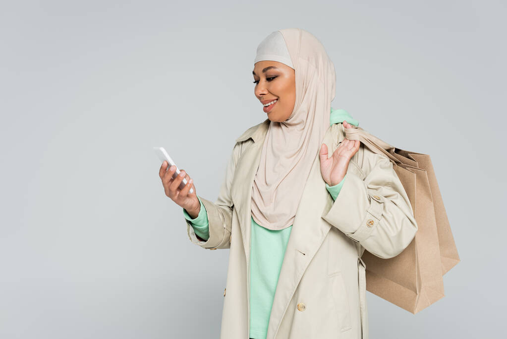 χαμογελαστή πολυφυλετική γυναίκα με καμπαρντίνα και μαντίλα κρατώντας τσάντες για ψώνια και κοιτάζοντας smartphone απομονωμένο σε γκρι - Φωτογραφία, εικόνα