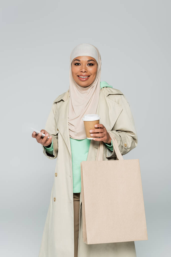 mujer musulmana multirracial de moda sosteniendo teléfono móvil y bebida para llevar con bolsa aislada en gris - Foto, imagen