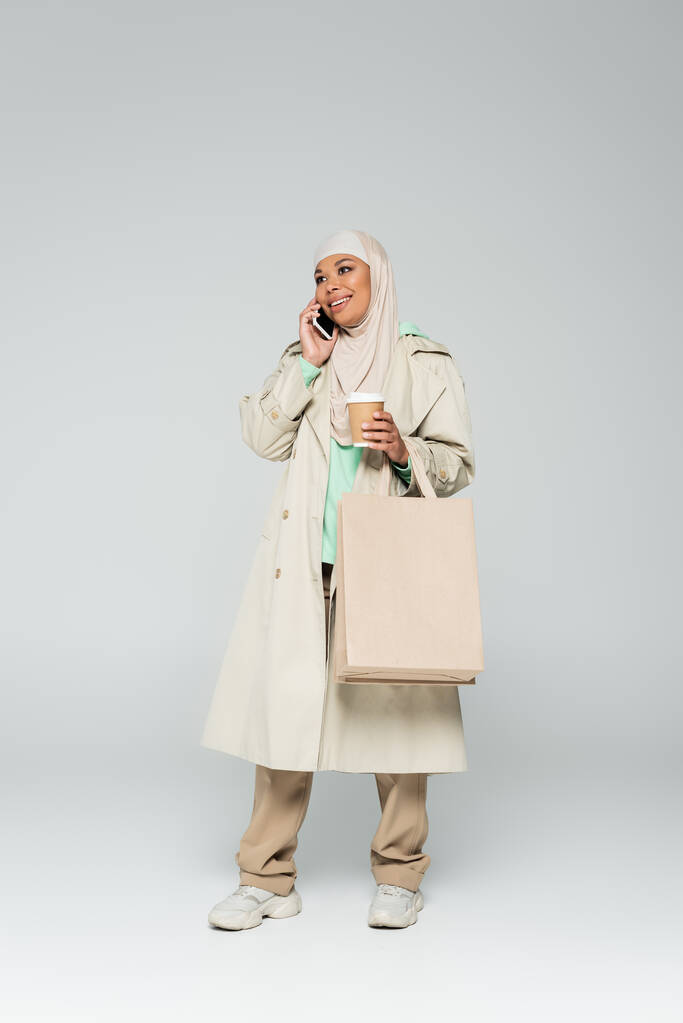 plná délka radostné multiraciální muslimka s kávou jít a nákupní tašky mluvit na smartphone na šedé  - Fotografie, Obrázek