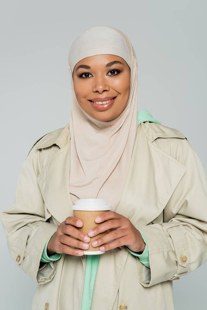 молодая многорасовая женщина в хиджабе и стильном плаще держа на вынос напиток и улыбаясь в камеру изолированы на серый - Фото, изображение