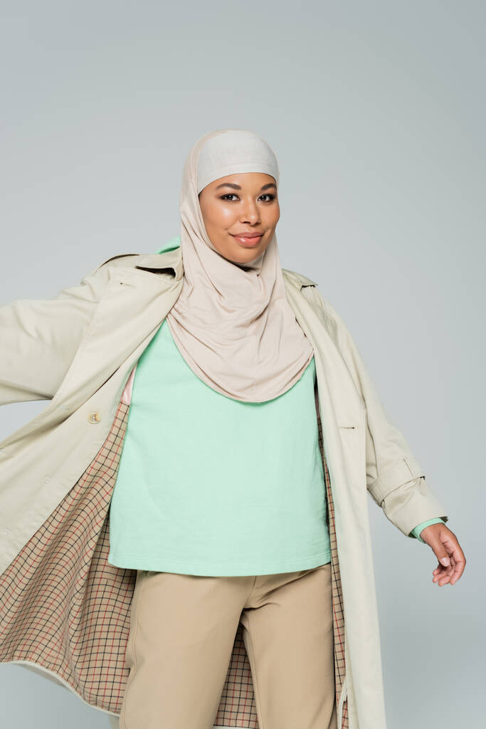 carefree multiracial woman in muslim hijab and trench coat posing and smiling at camera isolated on grey - Photo, Image