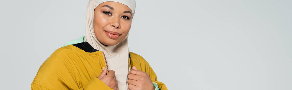 alegre mujer musulmana multirracial en hijab y chaqueta con estilo amarillo mirando la cámara aislada en gris, pancarta - Foto, Imagen