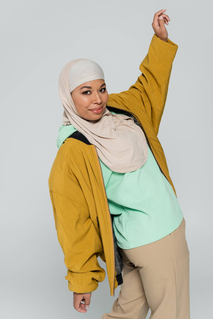 módní multiraciální žena ve žluté bombardér bunda a hidžáb pózování se zvýšenou rukou izolované na šedé - Fotografie, Obrázek