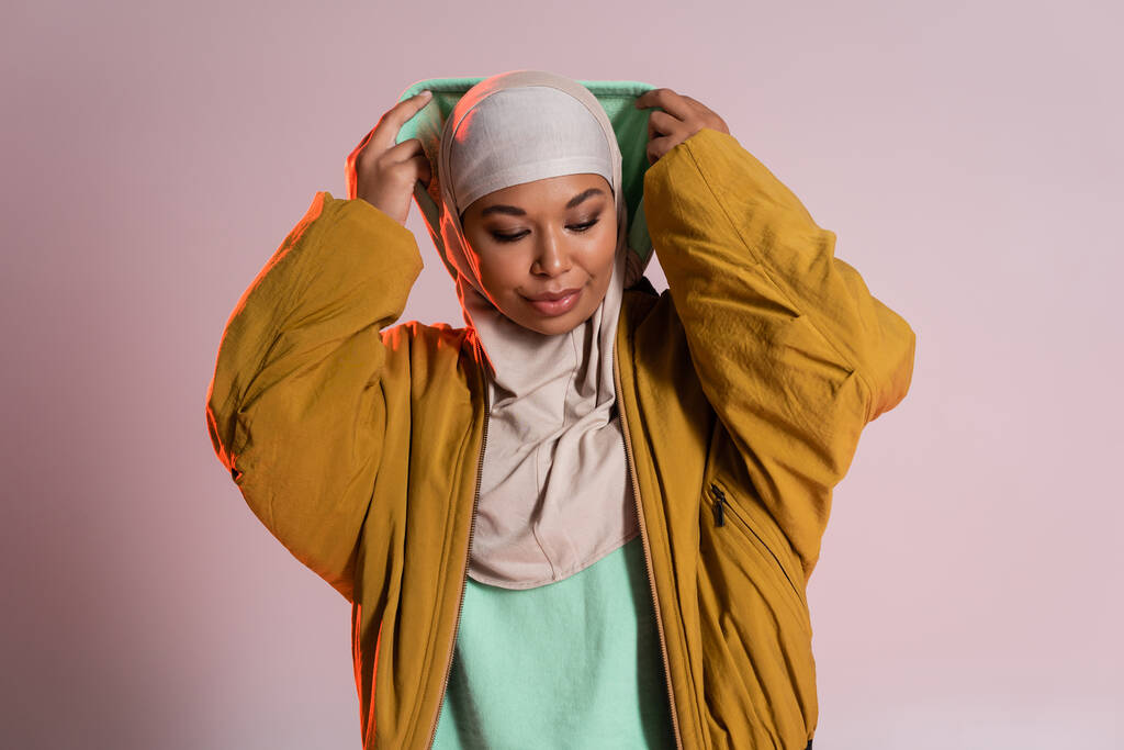 mujer joven y de moda multirracial en hijab y chaqueta bombardero amarillo con capucha sobre fondo gris rosado - Foto, Imagen