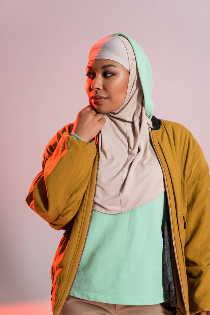молодая и красивая многорасовая женщина в желтой куртке бомбардировщика и хиджабе смотрит в сторону на сером и розовом фоне - Фото, изображение