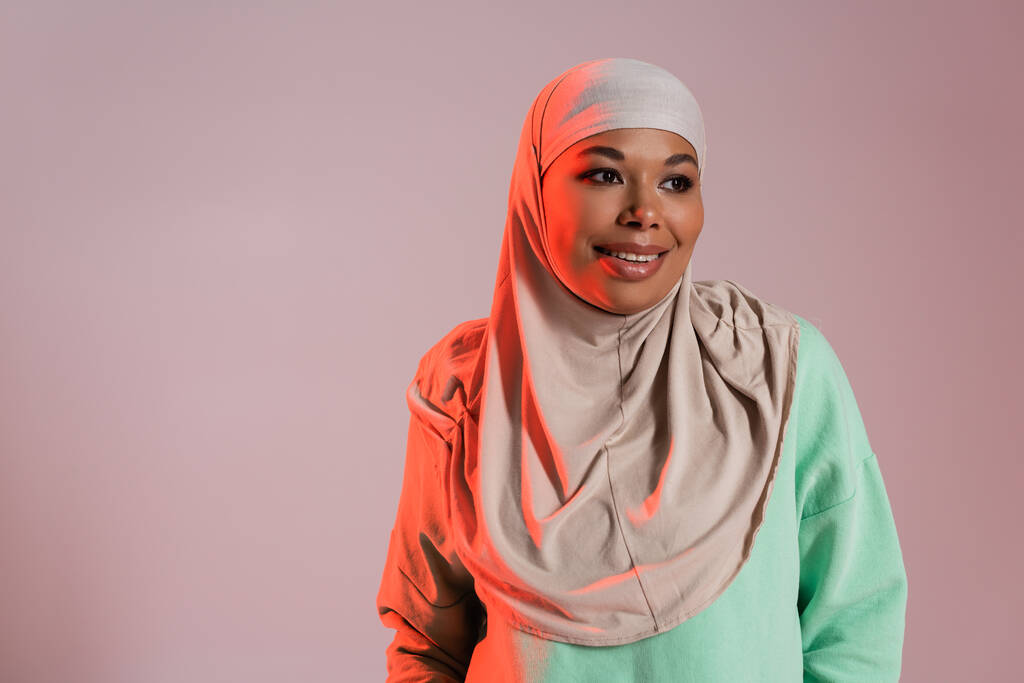 zorgeloze multiraciale moslim vrouw in traditionele hijab weg te kijken geïsoleerd op roze grijs - Foto, afbeelding
