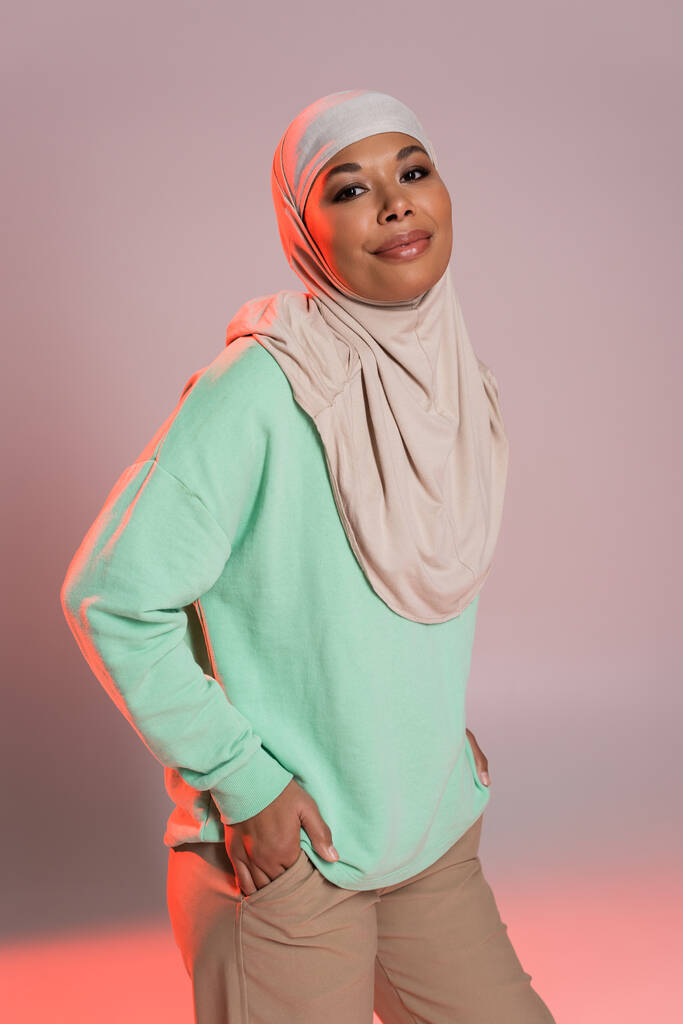 heureuse femme multiraciale en hijab et chemise verte à manches longues posant les mains dans les poches sur fond gris et rose - Photo, image