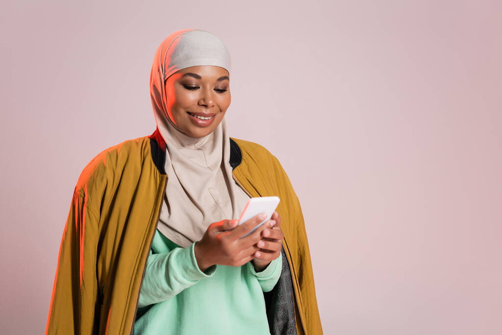 mujer multirracial complacido en chaqueta bombardero amarillo y hijab utilizando el teléfono móvil aislado en gris rosado - Foto, imagen