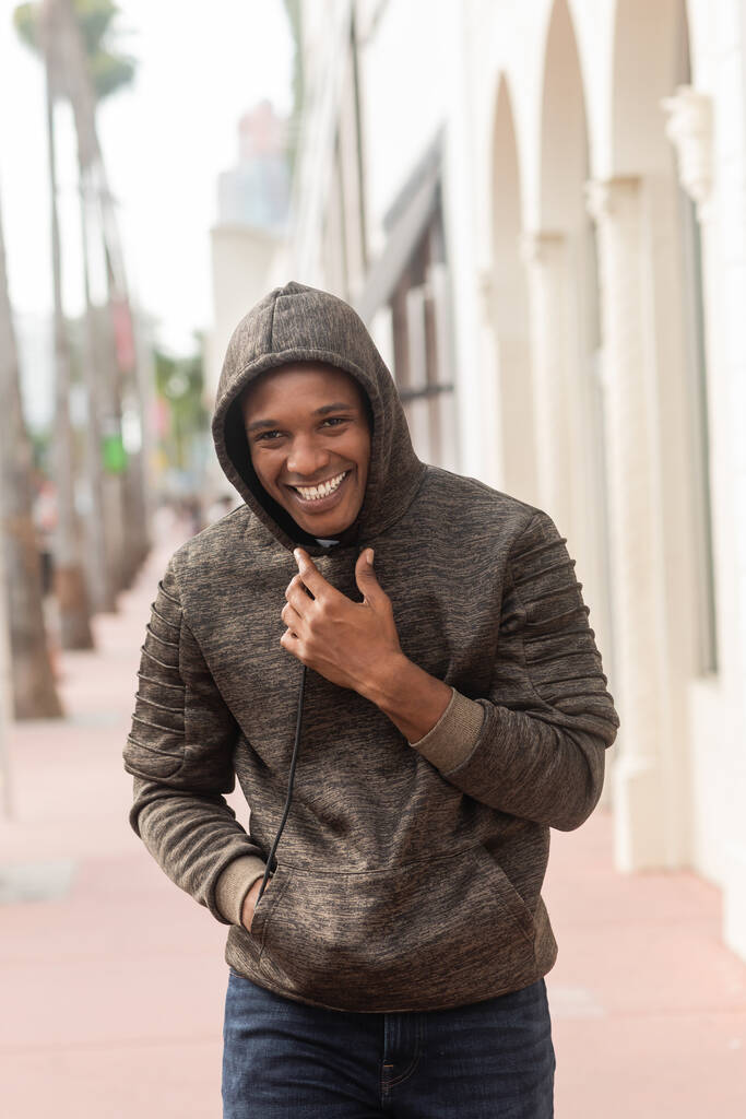 vreugdevolle Afrikaanse amerikaanse man in hoodie lopen met de hand in zak in Miami  - Foto, afbeelding