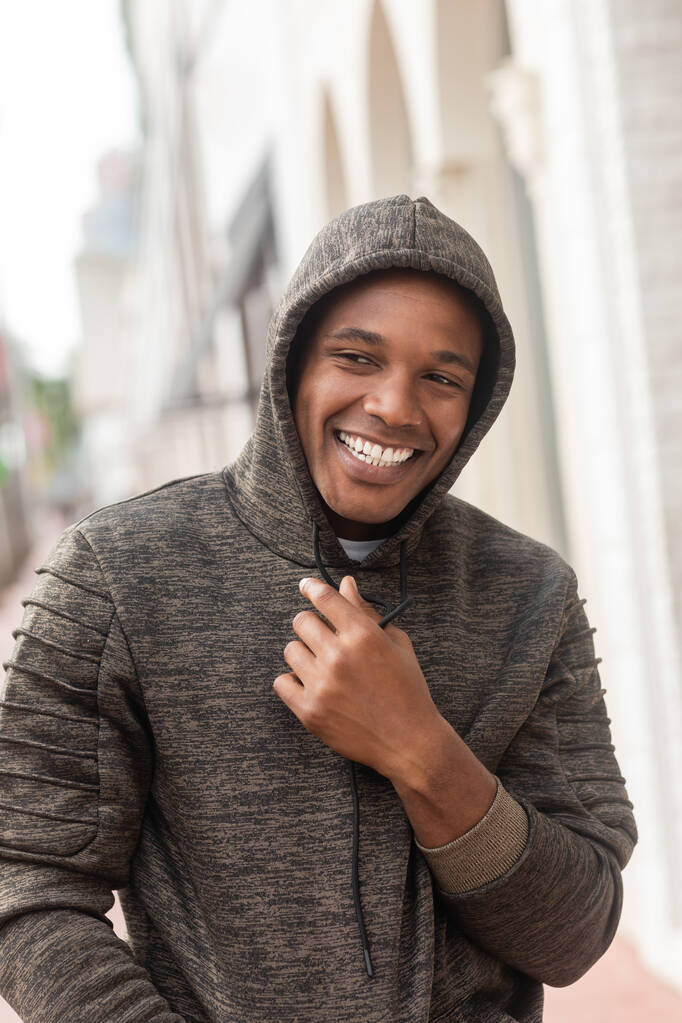 vrolijk Afrikaans amerikaanse man in capuchon sweatshirt weg te kijken terwijl glimlachen buiten  - Foto, afbeelding