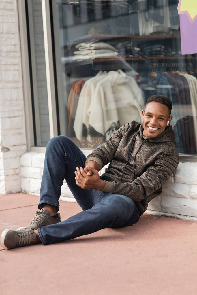 volledige lengte van vrolijke Afrikaanse Amerikaanse man in hoodie zitten in de buurt showcase van de winkel in Miami  - Foto, afbeelding