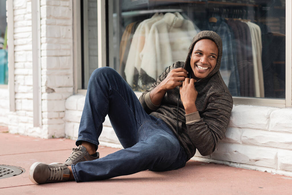 teljes hosszában boldog afro-amerikai férfi kapucnis pulóverben ül a kirakat közelében üzlet Miami  - Fotó, kép