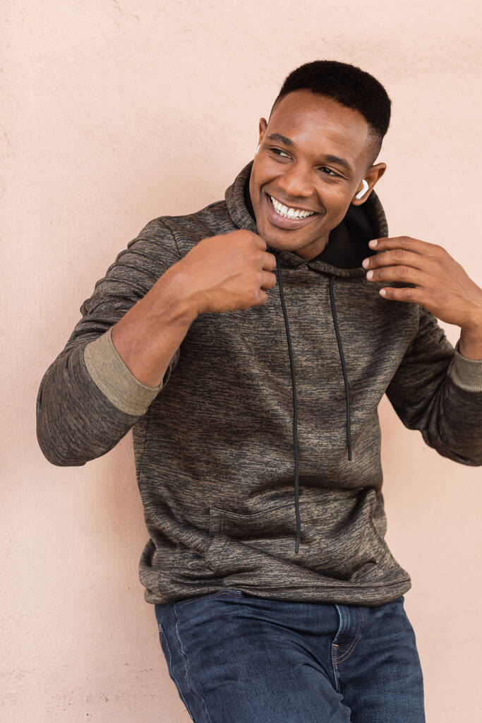 fröhlicher afrikanisch-amerikanischer Mann in drahtlosem Kopfhörer, der nahe der Wand Musik hört  - Foto, Bild