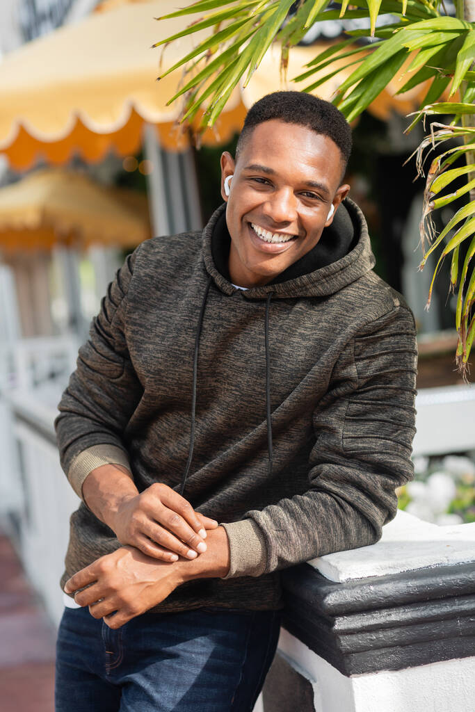 Unbekümmerter Afroamerikaner mit drahtlosen Kopfhörern, der draußen lächelt  - Foto, Bild