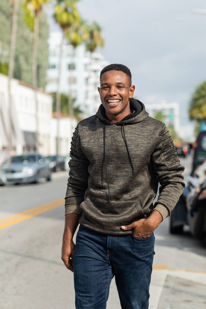 boldog afro-amerikai férfi kapucnis pulcsiban és farmerban, kéz a kézben sétál Miami utcáin.  - Fotó, kép