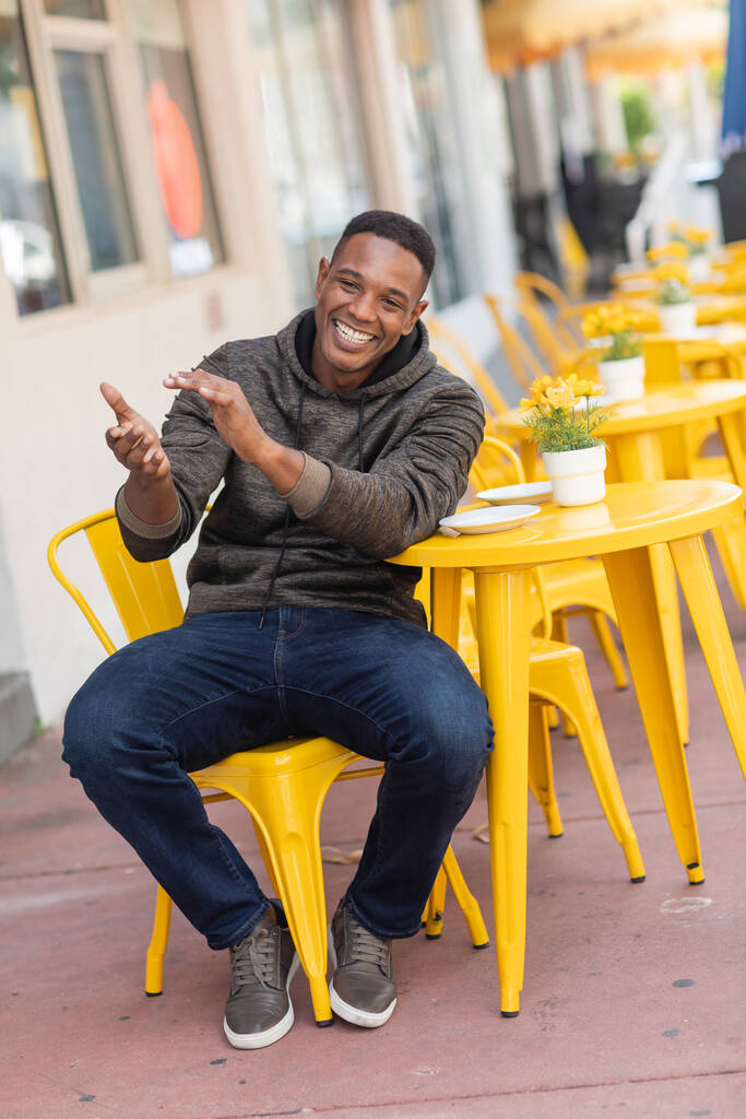 ξέγνοιαστος αφροαμερικάνος με κουκούλα και τζιν κάθεται στο τραπέζι του μπιστρό σε υπαίθριο καφέ  - Φωτογραφία, εικόνα
