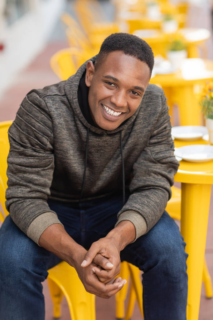 vreugdevolle Afrikaanse amerikaanse man in hoodie en jeans zitten aan bistro tafel in outdoor cafe  - Foto, afbeelding