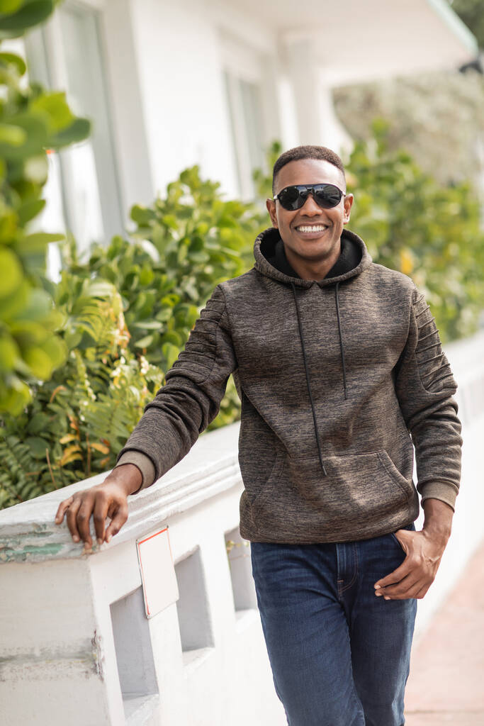 hombre afroamericano positivo en sudadera con capucha y gafas de sol con estilo de pie al aire libre  - Foto, Imagen