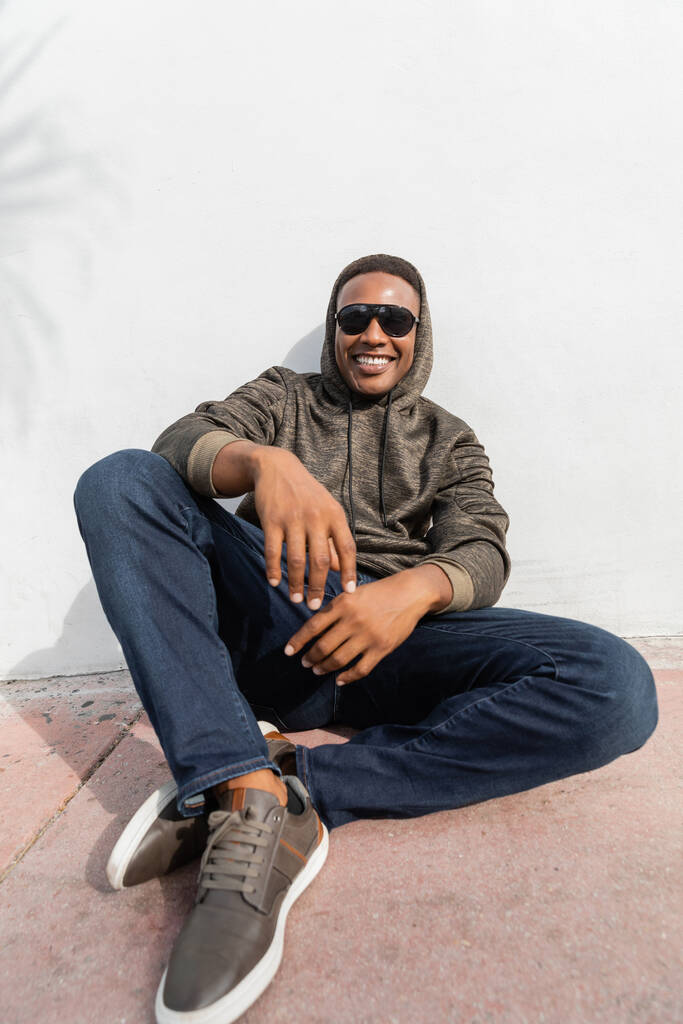 homem americano africano alegre em óculos de sol elegantes sentado perto da parede branca  - Foto, Imagem