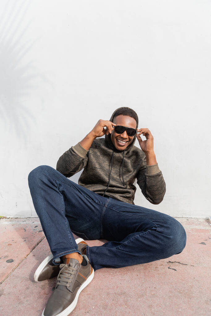 χαμογελαστός Αφροαμερικάνος άνδρας με κομψά γυαλιά ηλίου κάθεται κοντά σε λευκό τοίχο  - Φωτογραφία, εικόνα
