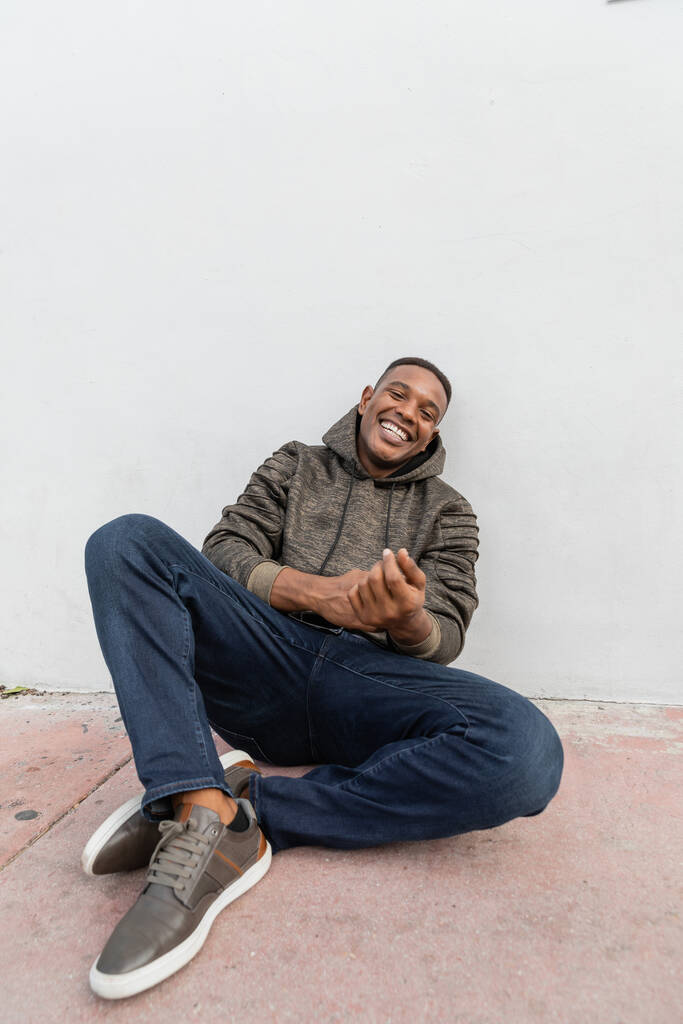 boldog afro-amerikai férfi farmerban és kapucnis mosoly mellett ülve fehér fal  - Fotó, kép