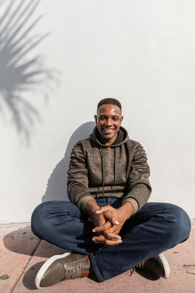 zorgeloos Afrikaans amerikaanse man in jeans en hoodie zitten in de buurt van witte muur  - Foto, afbeelding