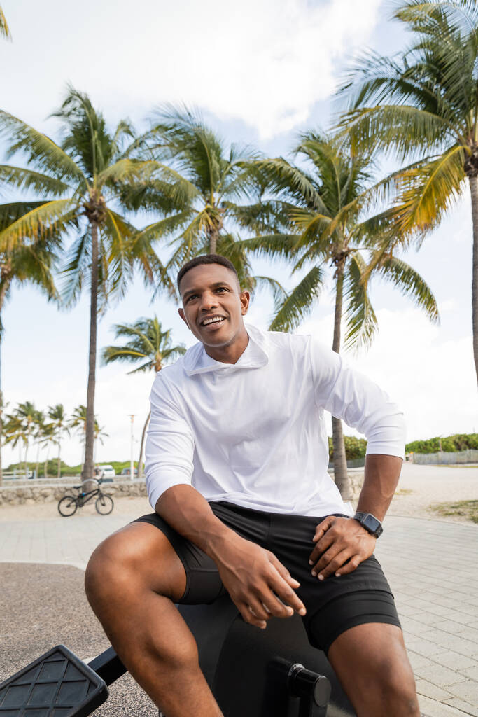 alegre homem americano africano sentado no equipamento cross trainer no ginásio ao ar livre em Miami - Foto, Imagem
