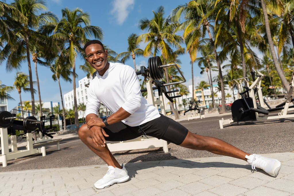 longitud completa de deportista afroamericano feliz estiramiento en el gimnasio al aire libre en Miami  - Foto, Imagen