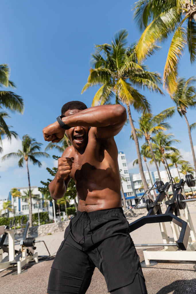 luchador afroamericano emocional y sin camisa en pantalones cortos haciendo ejercicio en Miami Beach  - Foto, imagen
