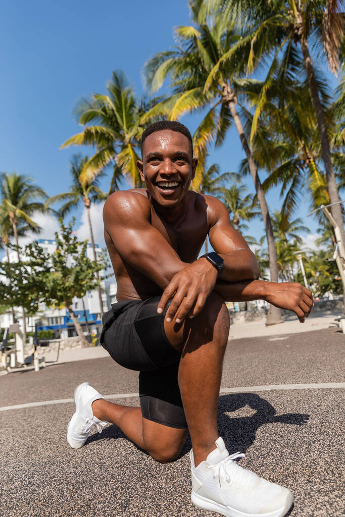 マイアミのヤシの木の隣でトレーニングした後微笑むシャツレスのアフリカ系アメリカ人スポーツマン - 写真・画像