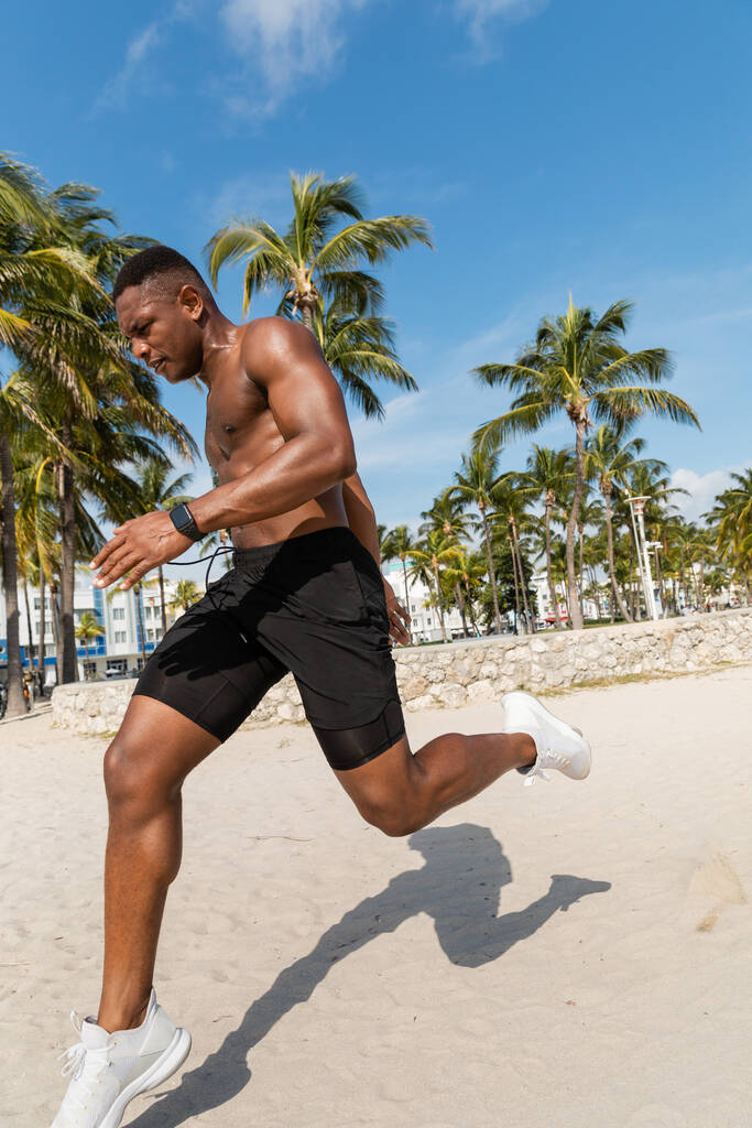 マイアミビーチのヤシの木の隣で砂の上を走っているショートパンツで筋肉質のアフリカ系アメリカ人男性 - 写真・画像