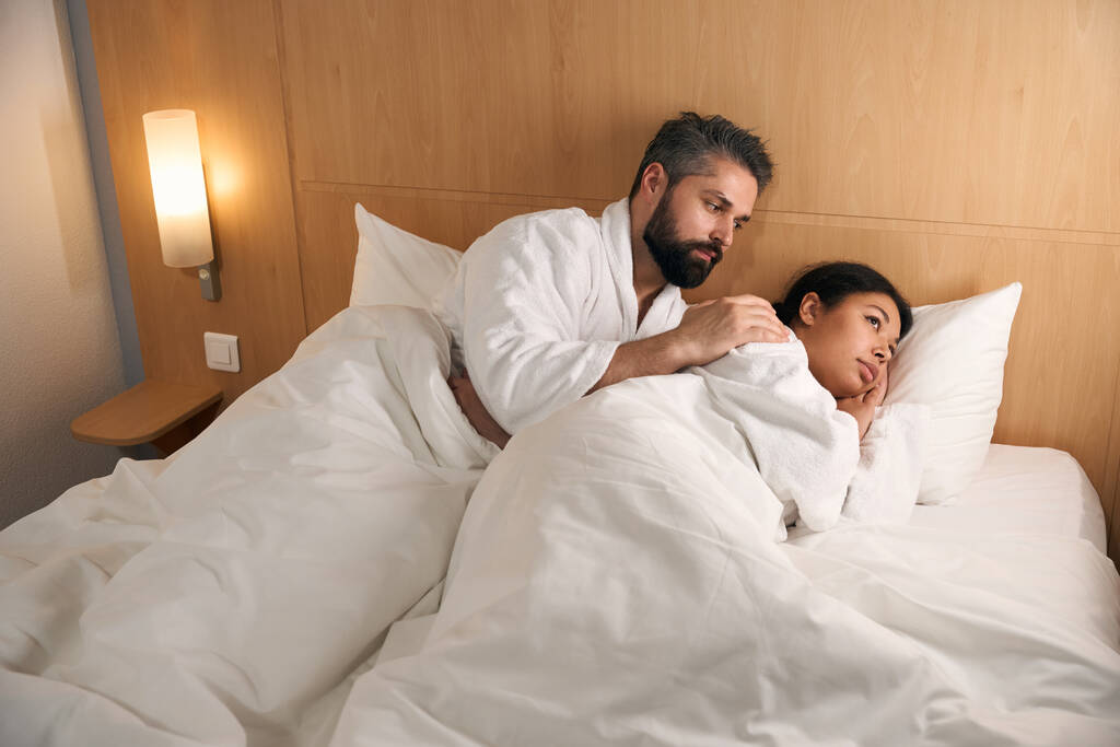 zorgzame man liggend in bed terwijl aanraken schouder van zijn low-spirited vriendin - Foto, afbeelding