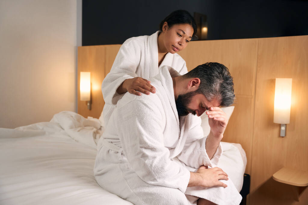 jonge vrouw kloppend op schouder verdrietig man gezeten op rand van bed - Foto, afbeelding