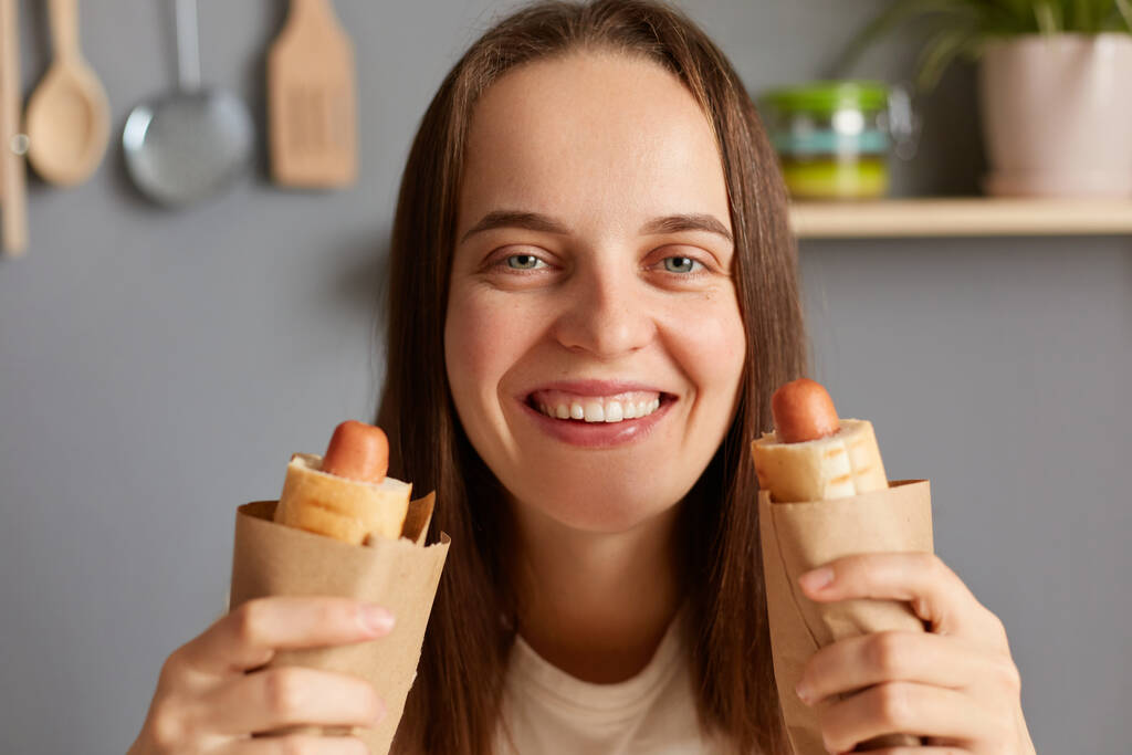 Nezdravé jídlo. Hnědé vlasy Kavkazský mladý dospělý pozitivní žena drží dva hot dogy cítí hlad, dívá se usmívá se do kamery, jíst nezdravé jídlo, těší ukončení stravy. - Fotografie, Obrázek