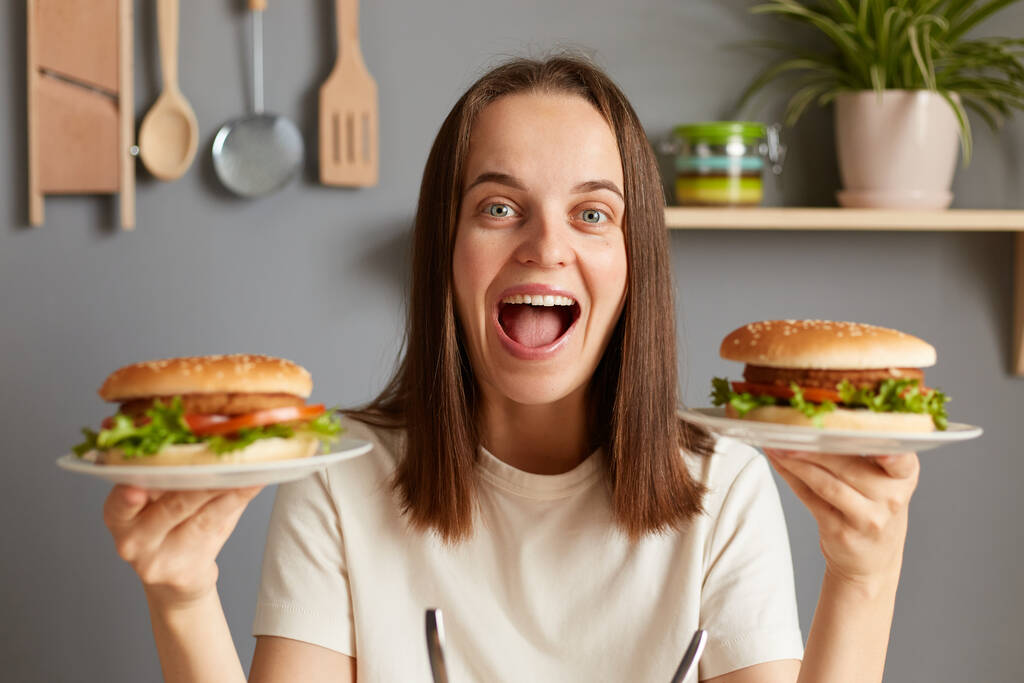 Indoor shot van verbaasd lachende vrouw met twee heerlijke verse hamburgers met sappige geroosterde kip en raketsalade, extreem hongerig, wow schreeuwen van geluk, genieten van junk food. - Foto, afbeelding