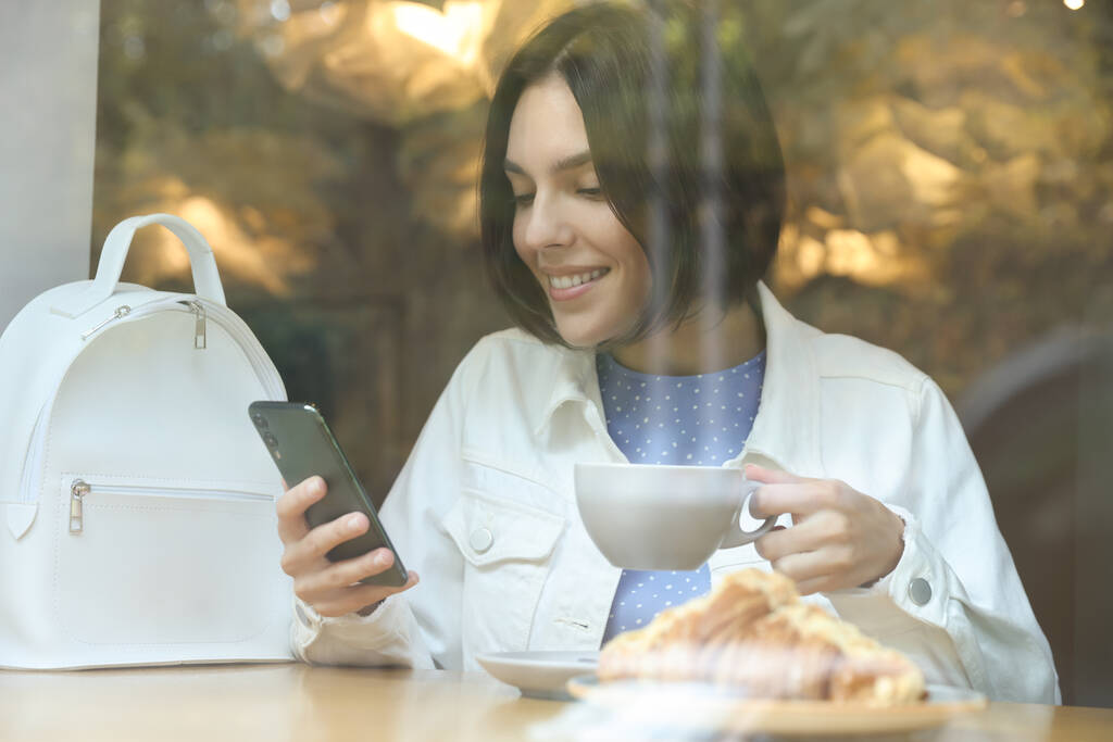 Promozione Speciale. Felice giovane donna con tazza di bevanda utilizzando smartphone in caffè, vista dall'esterno - Foto, immagini
