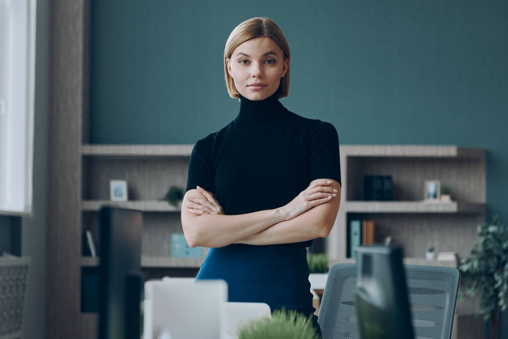 Selbstbewusste Geschäftsfrau blickt in die Kamera, während sie in der Nähe ihres Arbeitsplatzes im Büro steht - Foto, Bild