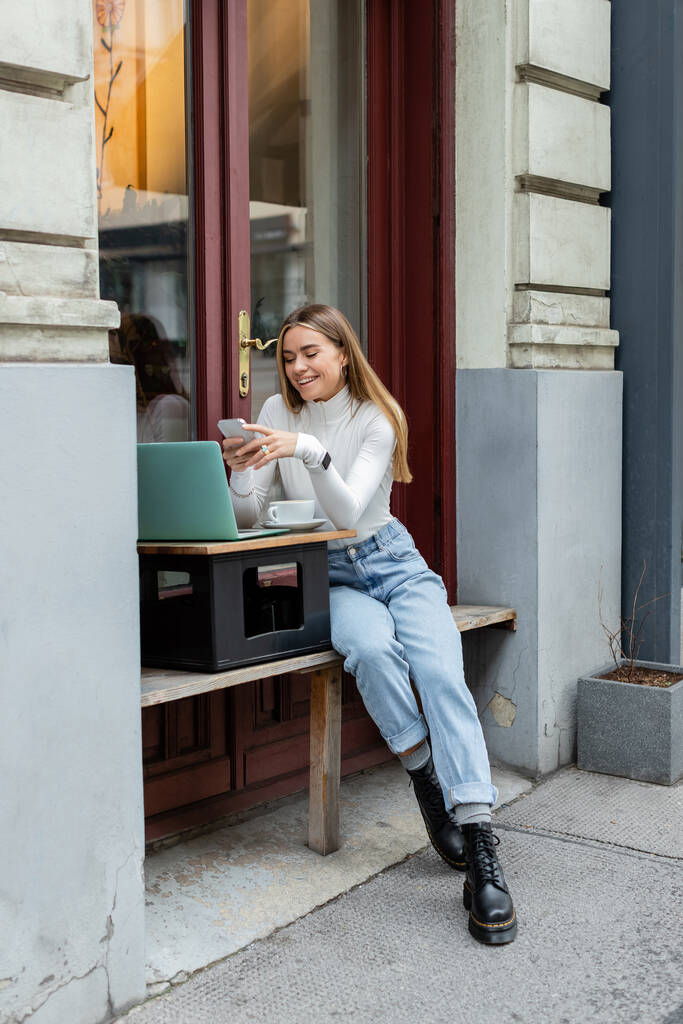 wesoła kobieta korzystająca ze smartfona w pobliżu laptopa i filiżanki kawy siedząc w kawiarni na ulicy w Wiedniu  - Zdjęcie, obraz