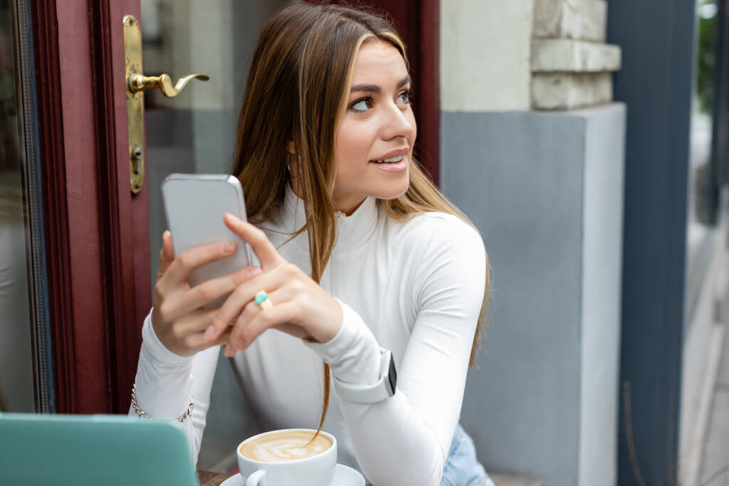 glimlachende jonge vrouw met smartphone in de buurt van een kopje cappuccino tijdens het zitten in cafe in Wenen  - Foto, afbeelding