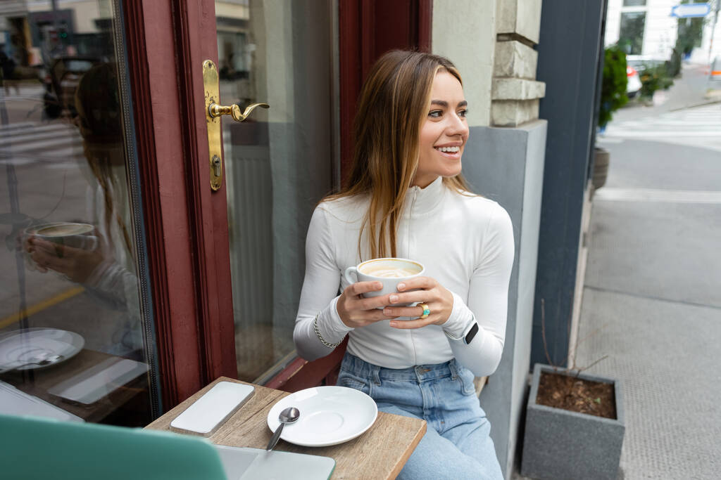 беззаботная женщина держит чашку капучино, сидя рядом с гаджетами на столе в кафе на улице в Вене  - Фото, изображение