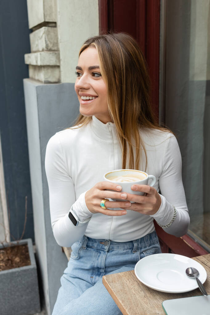 mulher despreocupada segurando xícara de latte enquanto sentado no café ao ar livre na rua em Viena  - Foto, Imagem