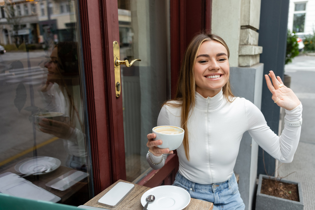 ξέγνοιαστη γυναίκα κρατώντας ένα φλιτζάνι καπουτσίνο ενώ χαιρετάει το χέρι σε καφέ στο δρόμο στη Βιέννη  - Φωτογραφία, εικόνα