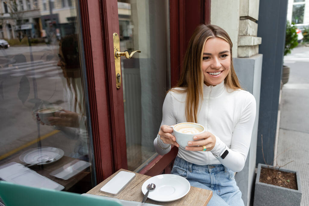 happy woman holding cup of cappuccino while sitting at bistro table with gadgets in cafe on street in Vienna  - Photo, Image