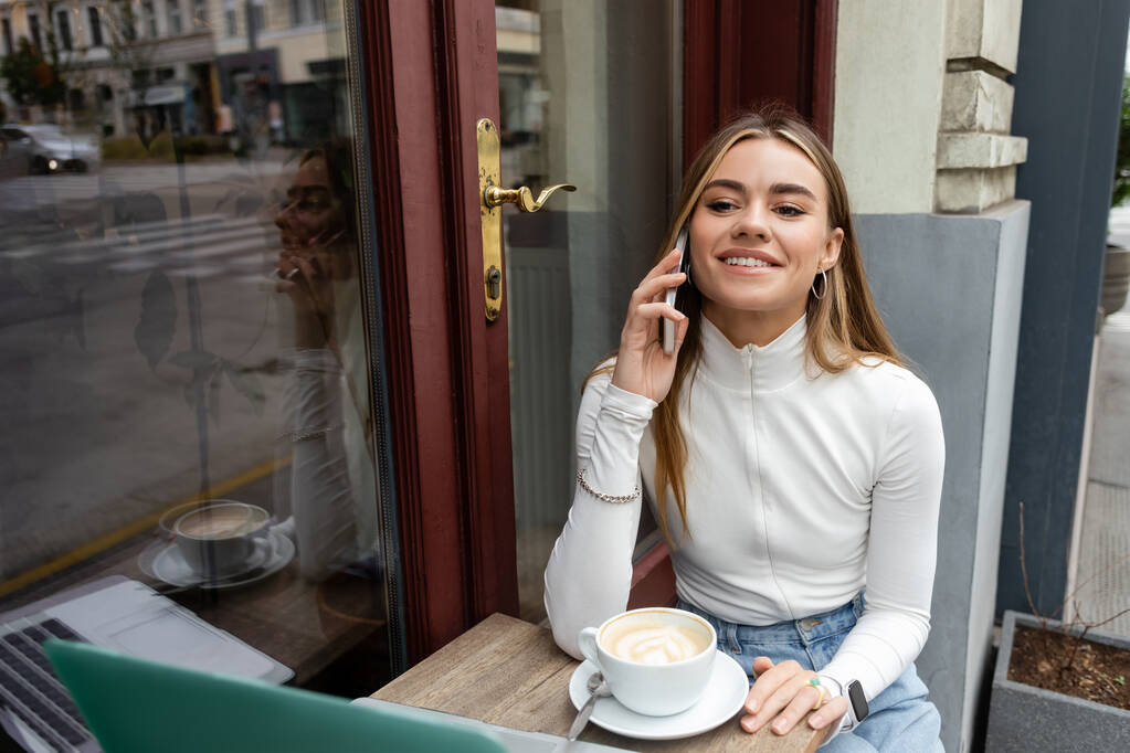 zadowolona kobieta rozmawia na smartfonie przy filiżance kawy siedząc w kawiarni na świeżym powietrzu w Wiedniu  - Zdjęcie, obraz