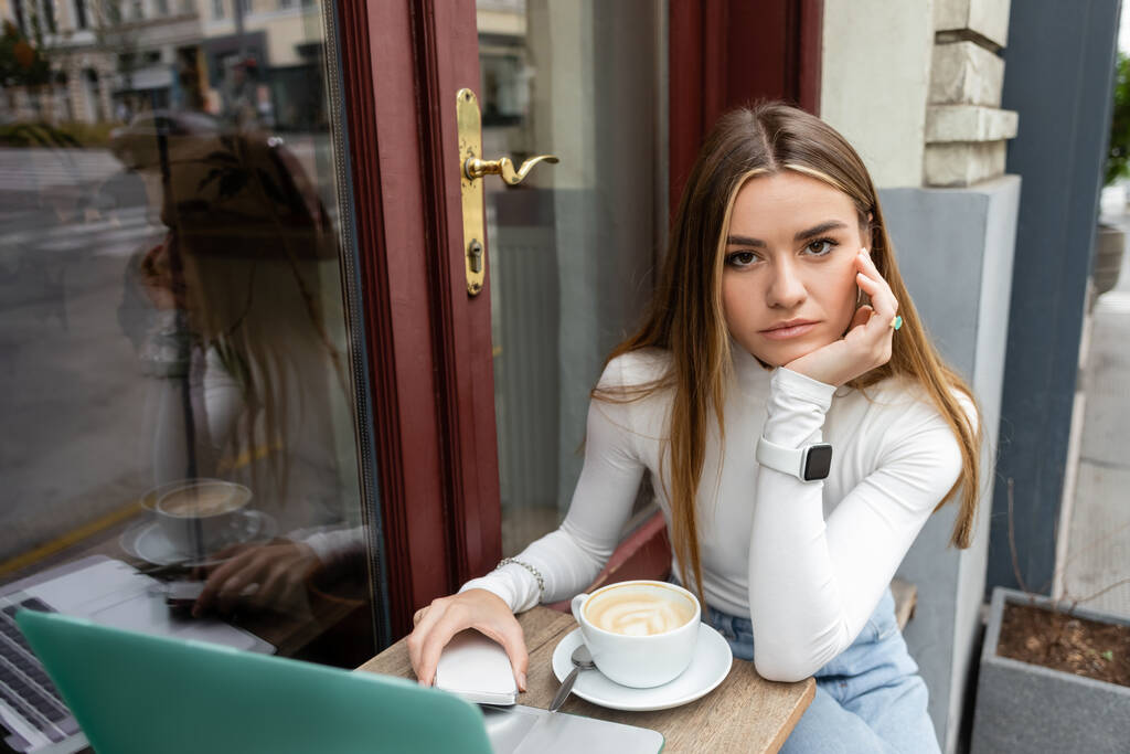 jonge vrouw houden smartphone in de buurt van kopje met cappuccino terwijl zitten aan bistro tafel in de buurt van laptop in cafe in Wenen  - Foto, afbeelding