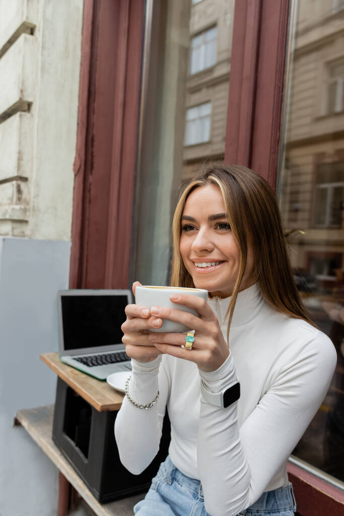 happy woman holding cup of coffee while sitting in outdoor cafe next to laptop with blank screen in Vienna  - Photo, Image