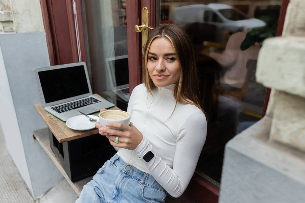 młoda kobieta trzyma filiżankę cappuccino siedząc w kawiarni na świeżym powietrzu obok laptopa z pustym ekranem w Wiedniu  - Zdjęcie, obraz