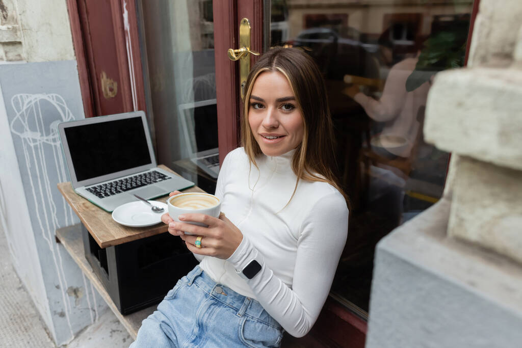 charming woman holding cup of cappuccino while sitting in outdoor cafe next to laptop with blank screen in Vienna  - Photo, Image