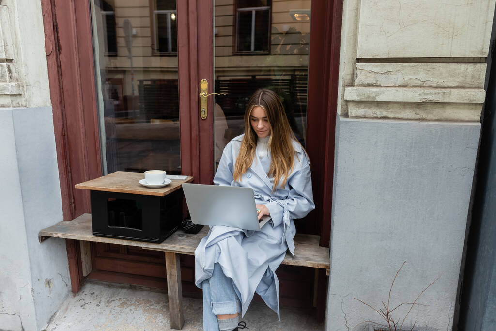 joven freelancer en gabardina azul usando laptop mientras está sentado al lado de una taza de café en una cafetería al aire libre en Viena   - Foto, Imagen