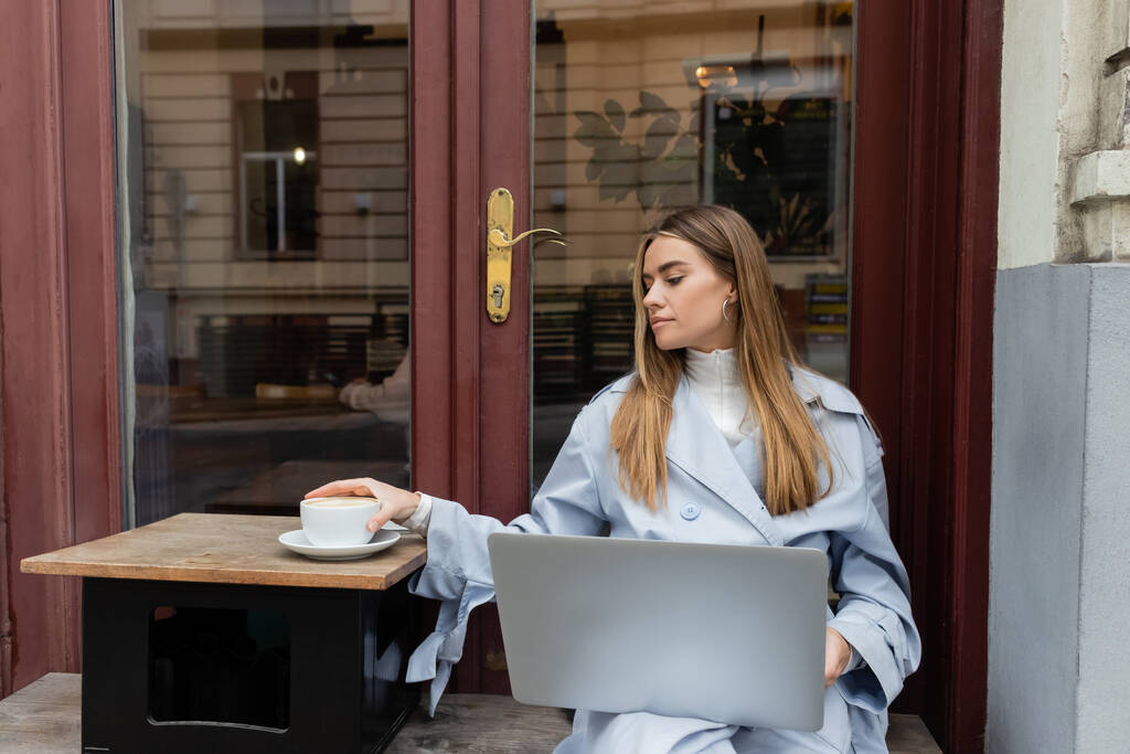 joven freelancer en gabardina azul usando portátil mientras llega a la taza de café en la cafetería al aire libre en Viena   - Foto, Imagen